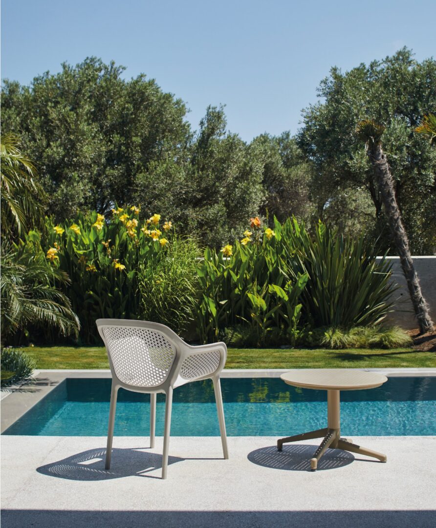 Contemporary gray outdoor commercial armchair with perforated back design placed beside a resort pool surrounded by lush Mediterranean landscaping and blue sky.