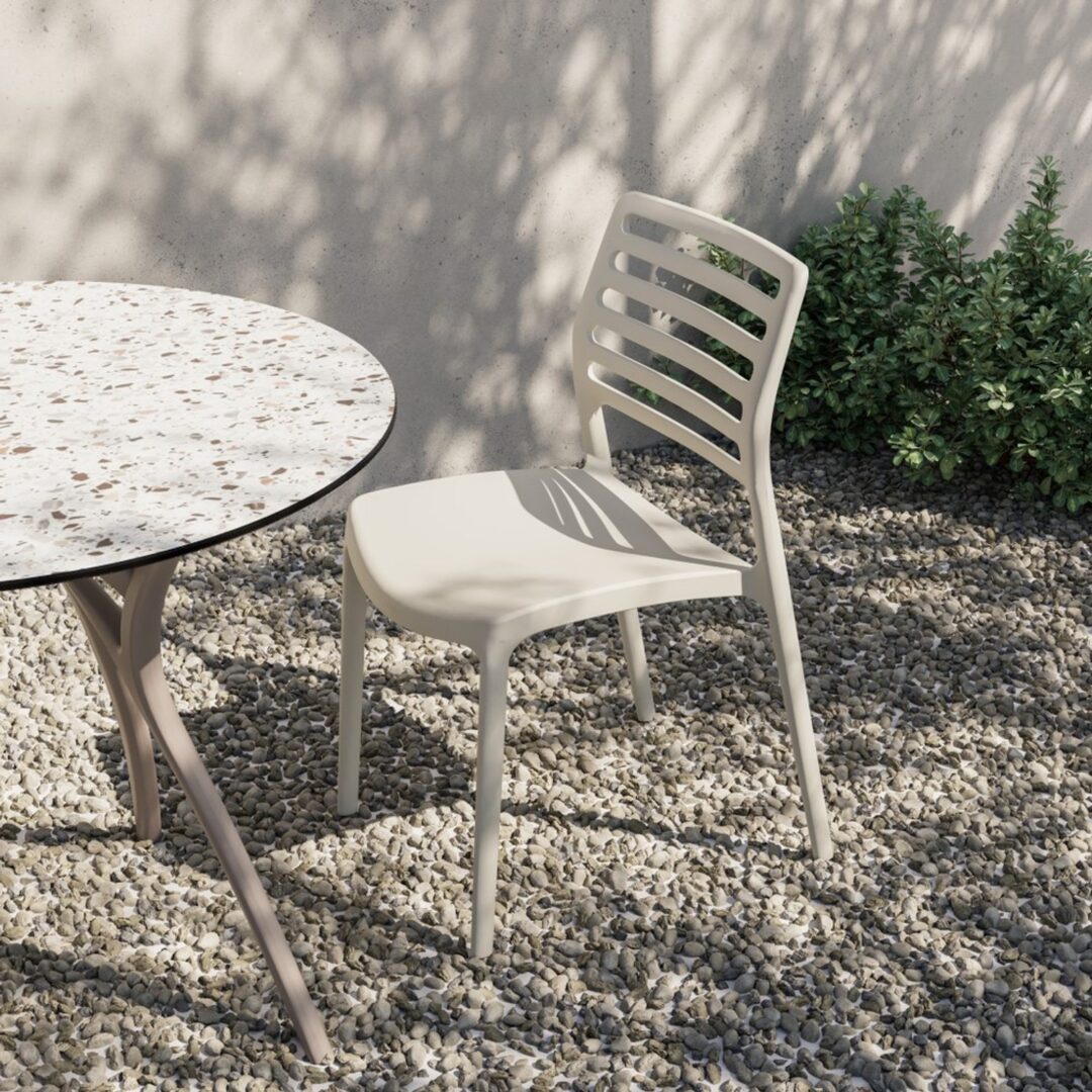 Ivory white Louise commercial outdoor chair with slatted back beside a terrazzo round table on a gravel garden surface with natural sunlight and soft wall shadow.
