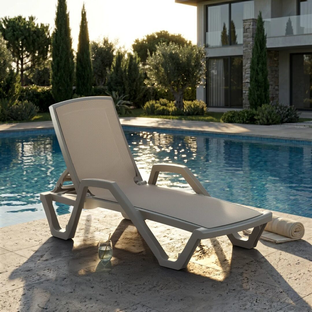 Premium mink and beige Mare commercial sun lounger with mesh sling seat and armrests positioned beside a luxury resort swimming pool at golden hour with cypress trees and stone villa.