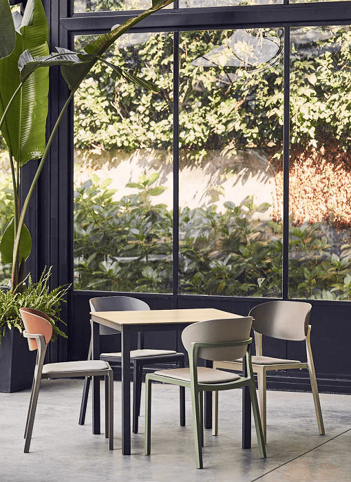 Architectural dining setting with mismatched commercial chairs in sage green, taupe, and blush tones surrounding a light wood table before floor-to-ceiling windows with lush garden views.